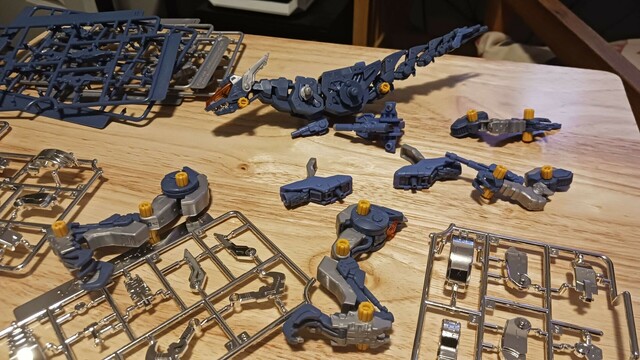 A photograph of the sprawl upon Beatrix's modeling desk. Abigail, the gun sniper (smallish blue raptor with some grey armour parts and a fancy tail, if you're not familiar) is picked apart into her component parts, the remaining parts of the B sprues sit stacked in the corner, and the three chrome sprues are spread across the rest of the space.