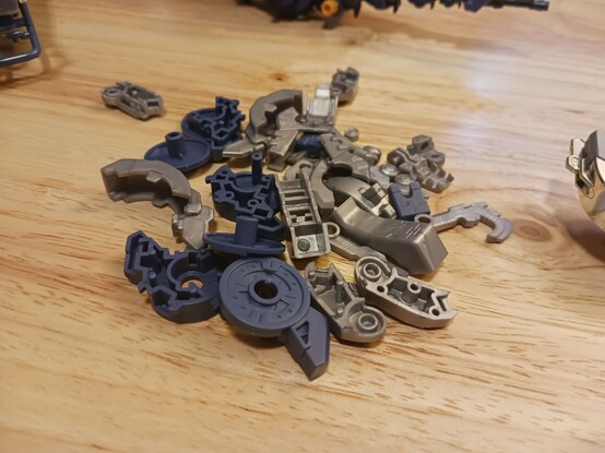 A photograph of a bunch of plastic parts in a pile on the desk. Mostly they're the grey parts which have been switched out for silver, but the hip pieces which were erroneously blue before are also here, swapped for the proper grey parts on the model. There's a few more parts, but I cleared them off before deciding to just pile everything up partway through the process.

They're in a bag in the original box now. No sense wasting them, just in case anything breaks in the future!