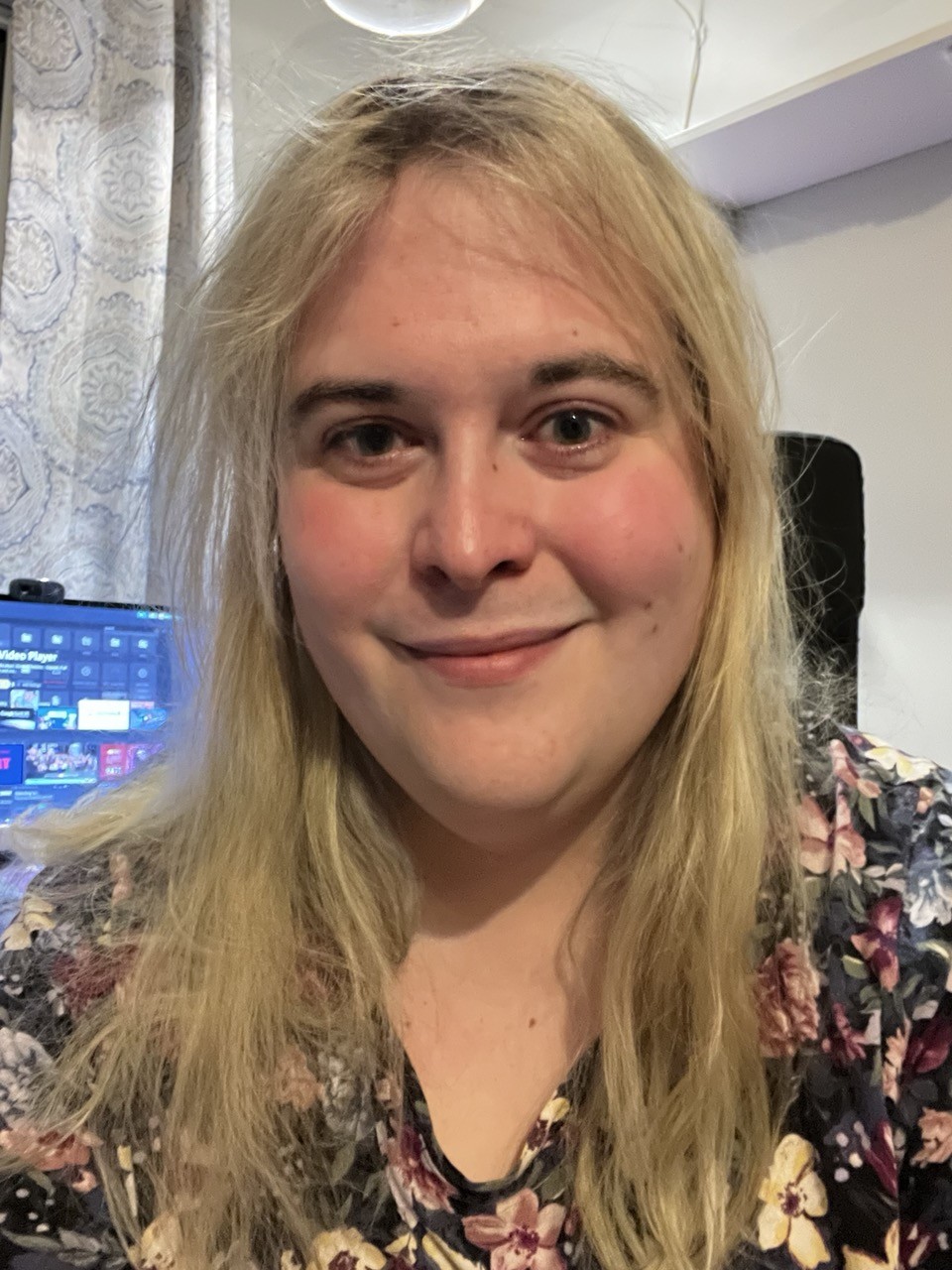 selfie of me, a white woman. i’m wearing a floral pattern shirt with burgundy, navy, and cream flowers dense on a black background. my blonde hair is framing my face. i am not wearing glasses. behind me is visible a shelf, a computer monitor, and a curtain. The lighting is warm