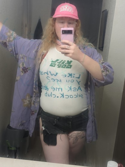 A mirror selfie of me, priestess of Shrek, wearing very short (how short? Well, I could easily get fingered wearing them. Tee hee :3) black denim cutoff shorts with a rip all the way down one of the side seams, a tight white form fitting tank top, that hugs my curves, with "kill cops" screen printed I'm green at the top, under that is a handwritten message in teal sharpie that reads "Like what you see? Ask me about girlcock.club." over that I'm wearing an oversized lavender hawaiian-style button down shirt. A little bit of my red bra strap is also peaking out. Atop my head is my signature hot pink "princess" ballcap and a pink and baby blue rhinestone studded collar.