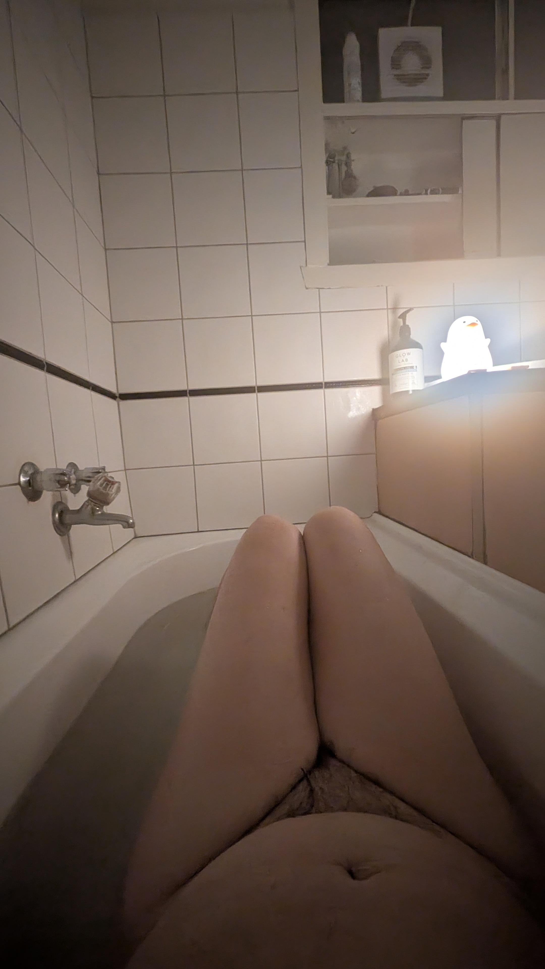 Looking down a naked person in a bathtub from just above their navel over their bent legs with knees jutting from the water in order to better fit. A glowing novelty duck shaped light illuminates the scene